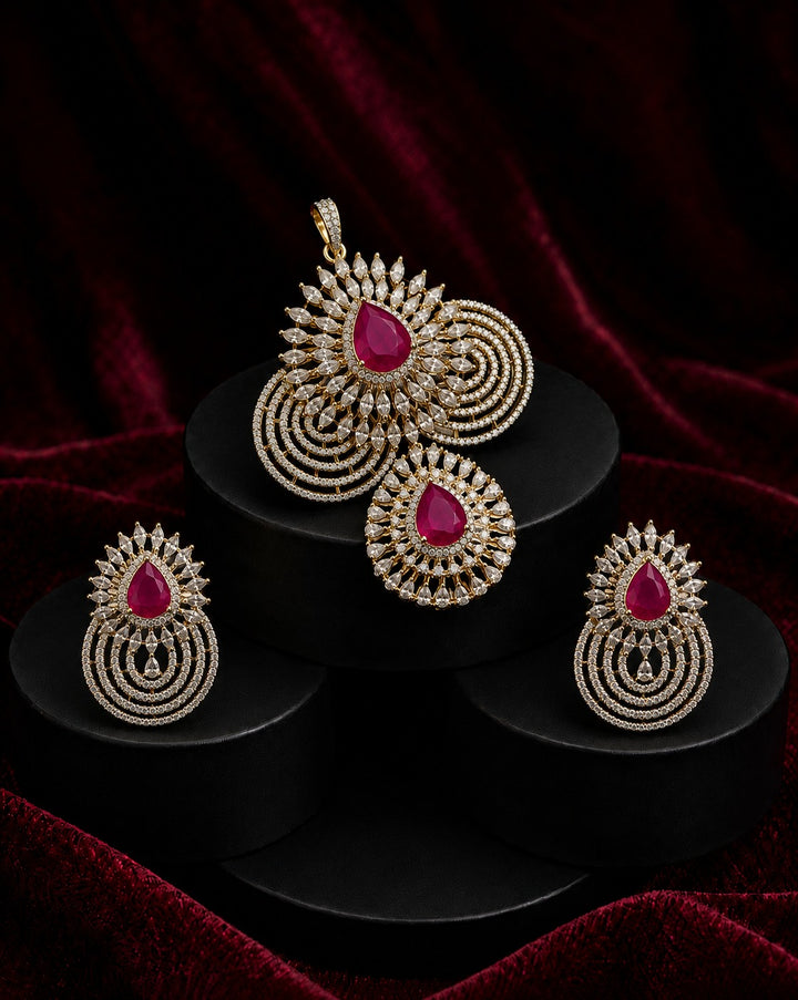 Gold-toned traditional Indian jewelry set featuring a pendant, earrings, and a large brooch, each adorned with a central teardrop-shaped pink gemstone and intricate circular gold pavé detailing, displayed on black pedestals against a dark red velvet background.