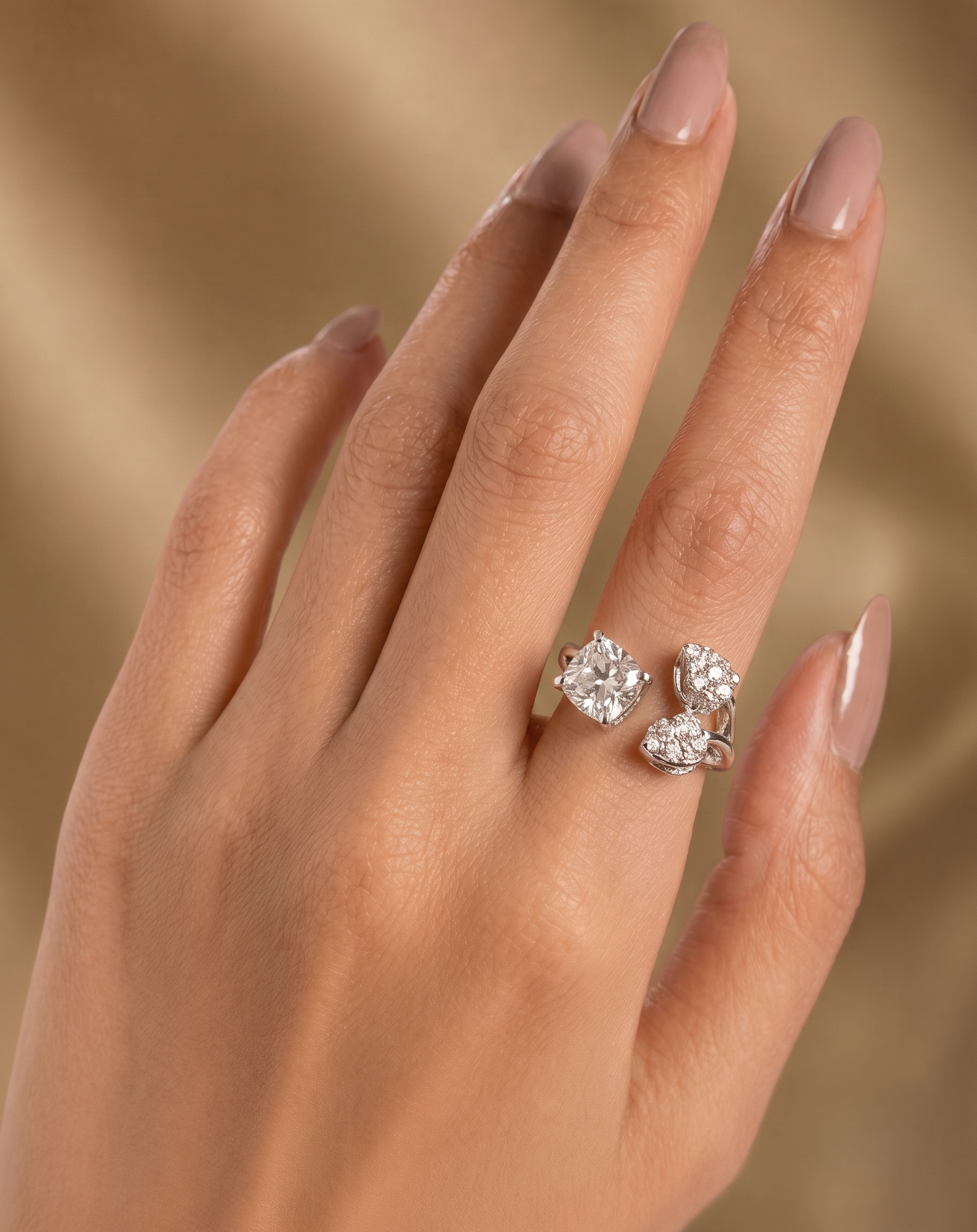 A close-up of a hand with manicured nails wearing a silver ring set with three clear round-cut gemstones, set against a soft, warm beige background.