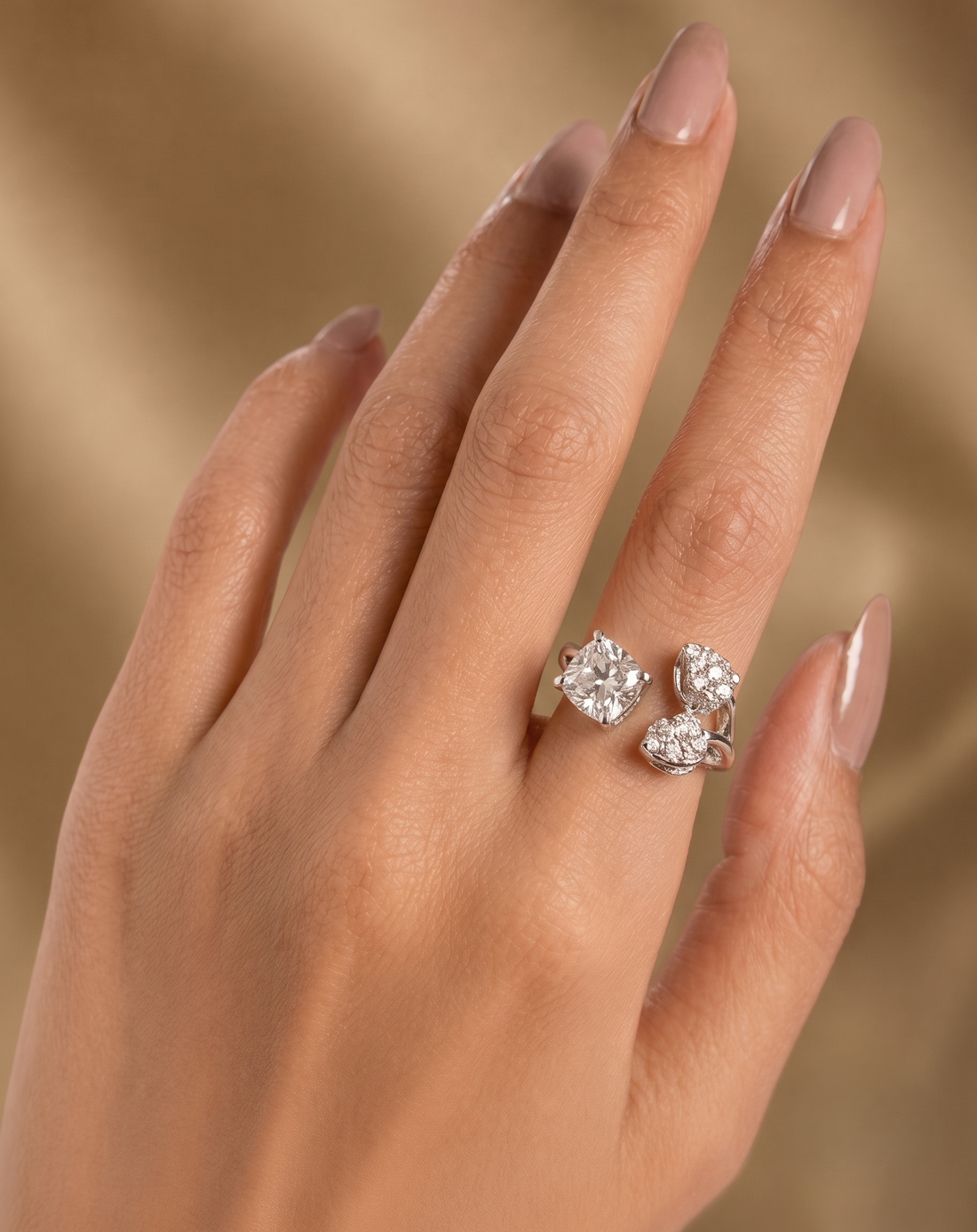 A close-up of a hand with manicured nails wearing a silver ring set with three clear round-cut gemstones, set against a soft, warm beige background.