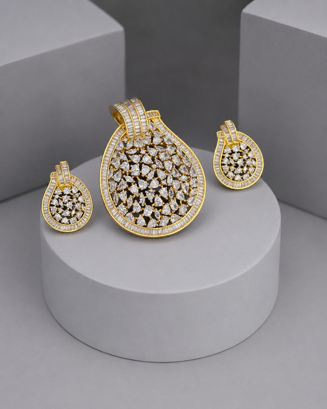 Gold jewelry set featuring a teardrop-shaped pendant and two matching drop earrings, all heavily embellished with clear stones, displayed on a light gray cylindrical pedestal.