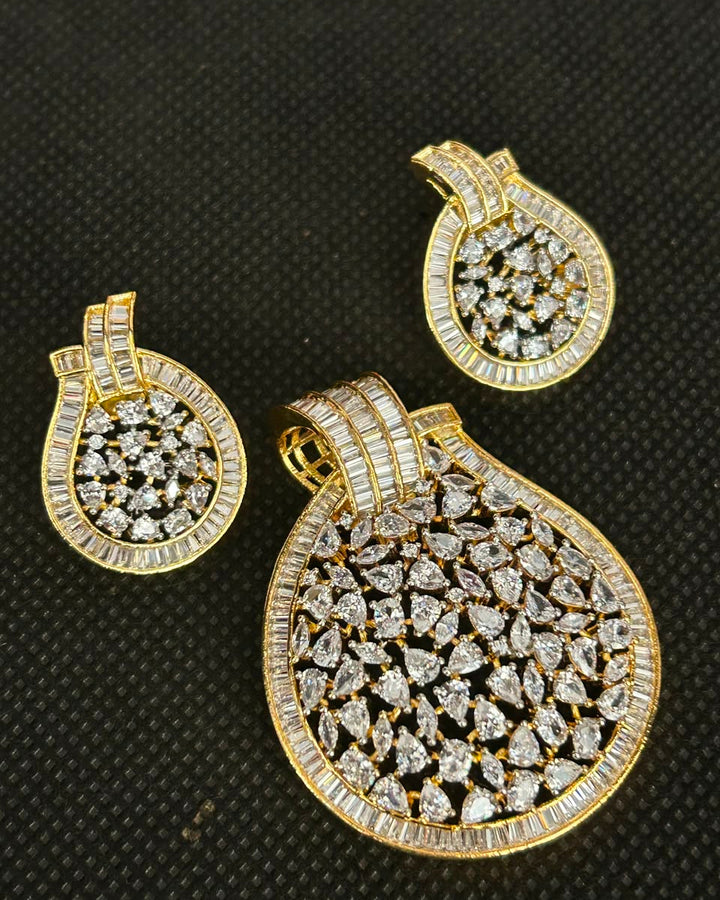 A gold jewelry set featuring a large teardrop-shaped pendant and two matching teardrop-shaped earrings, all heavily embellished with clear pavé gemstones, displayed on a black textured background.